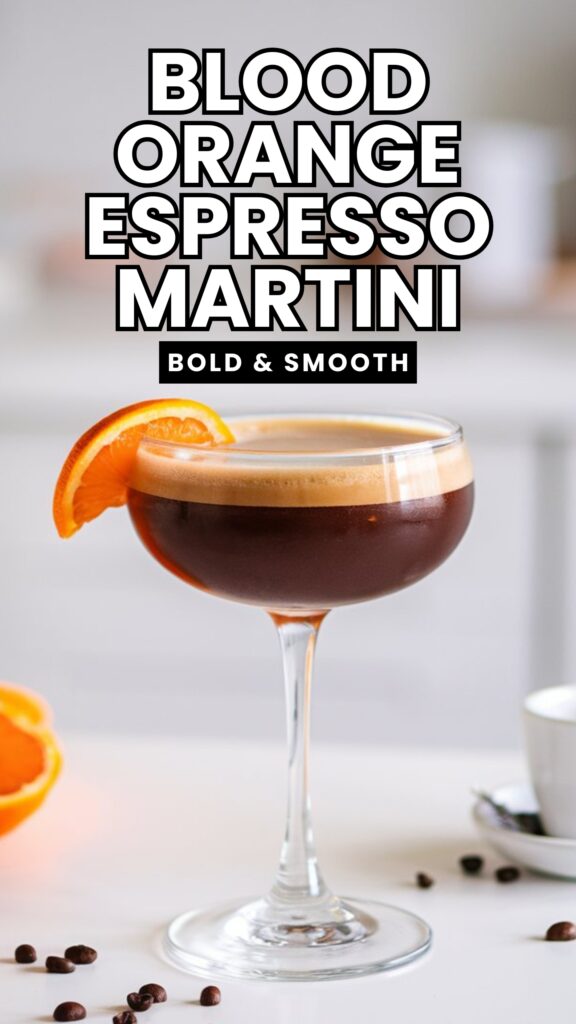 A glass of Blood Orange Espresso Martini with a foamy top and an orange twist garnish, set against a bright, minimal white kitchen with a white countertop. The drink has a deep, rich coffee color with a hint of citrus. A small espresso cup and coffee beans sit nearby for a stylish touch.