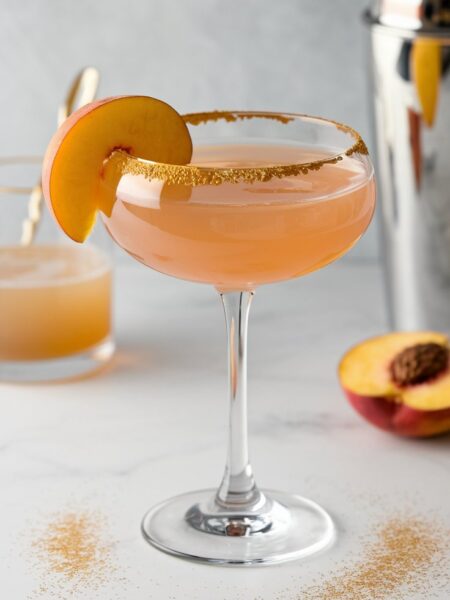 A refreshing peach martini cocktail in a martini glass garnished with fresh peach slices, sitting on a white marble countertop