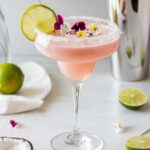 A creamy and vibrant Pink Coconut Margarita served in a classic margarita glass with a coconut-rimmed edge. The blush pink cocktail is garnished with a lime wheel and colorful edible flowers, creating a tropical and elegant presentation. Fresh limes, coconut flakes, and a cocktail shaker are in the background, enhancing the vacation-like atmosphere.