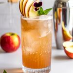 A bubbly Spiced Ginger Beer and Apple Fizz in a clear glass, filled with ice and topped with fresh apple juice and ginger beer. The drink has a warm golden hue and is garnished with thin apple slices, a cinnamon stick, fresh mint, and an edible flower. A cocktail shaker and fresh apples are in the background, creating a cozy, autumn-inspired scene.