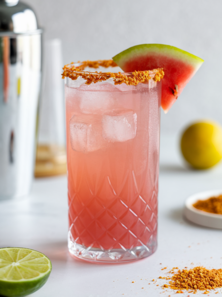 A vibrant pink Spicy Watermelon Paloma served in a Tajín-rimmed glass, garnished with a fresh watermelon slice, with condensation on the glass for a refreshing summer feel.