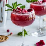 A romantic and refreshing Zero-Sugar Love Potion Punch served in an elegant coupe glass. The deep red mocktail is topped with fresh mint, frozen cranberries, and delicate rose petals, creating a festive Valentine's Day drink. A cocktail shaker and scattered rose petals add to the dreamy ambiance in the background.