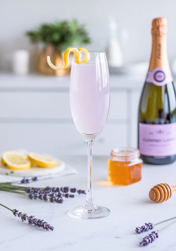 A beautifully crafted Lavender Honey French 75 served in a tall, chilled champagne flute with a delicate lemon twist garnish. The drink has a pale golden hue with a slight lavender undertone. The background features a bright, minimal white kitchen with fresh lavender sprigs, a honey jar, and a bottle of champagne to showcase the preparation process.