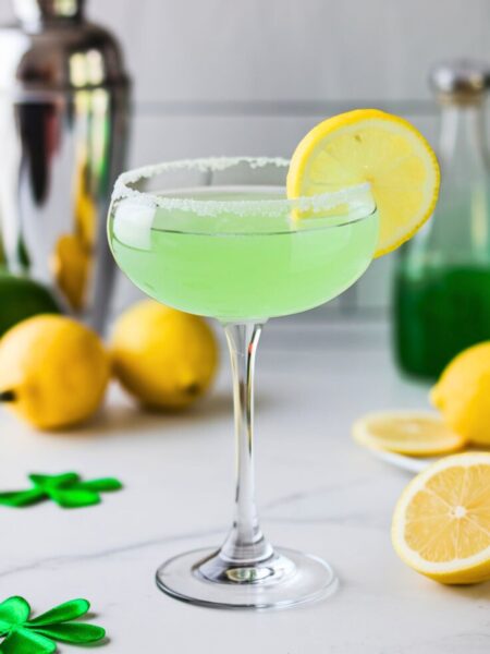 A Lucky Lemon Drop Martini served in a chilled martini glass with a sugar rim. The drink is a bright, citrusy green and garnished with a fresh lemon slice. The background is a clean white kitchen with a cocktail shaker and fresh lemons.