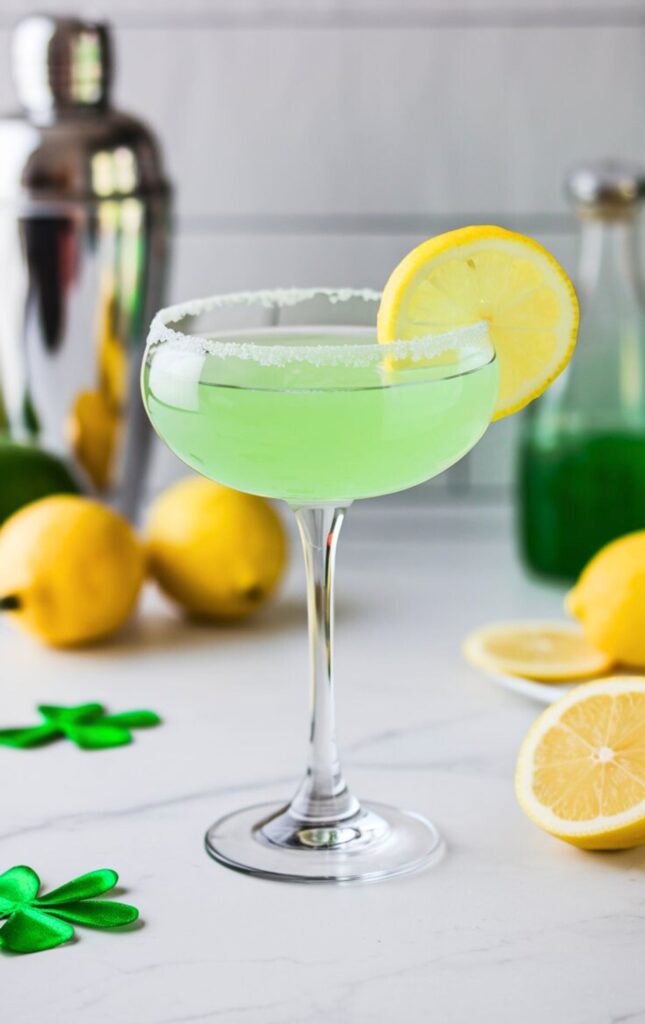 A Lucky Lemon Drop Martini served in a chilled martini glass with a sugar rim. The drink is a bright, citrusy green and garnished with a fresh lemon slice. The background is a clean white kitchen with a cocktail shaker and fresh lemons.