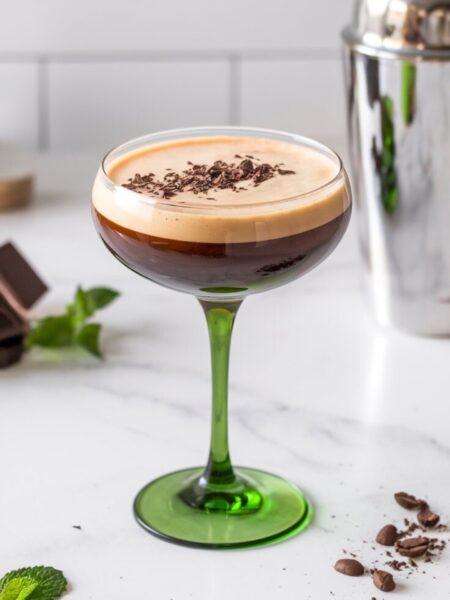 A rich and creamy Irish Cream Espresso Martini served in a chilled martini glass with a frothy top. The cocktail has a slight green tint from the crème de menthe, dusted with chocolate shavings and espresso beans. The background is a bright, clean, minimal white kitchen with a white countertop, evoking a cozy yet festive St. Patrick’s Day vibe. A hint of mint green is visible in the drink, reinforcing the refreshing minty flavor.