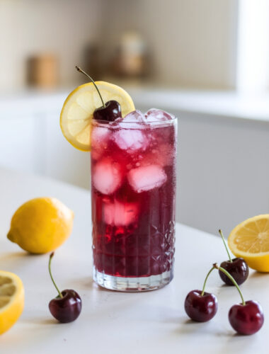 A refreshing glass of Cortisol-Balancing Tart Cherry Spritz with ice, garnished with a cherry and a lemon slice. The drink has a deep red hue with a slight effervescent glow.