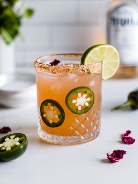 A Spicy Rose Margarita served in a salt-rimmed rocks glass with a lime wedge and dried rose petal garnish. The drink has a golden hue with jalapeño slices floating inside.