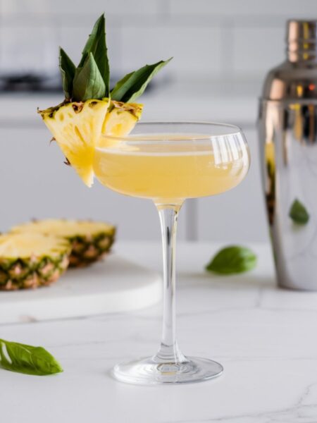 A bright and refreshing Pineapple Basil Martini served in a chilled martini glass, garnished with a fresh basil leaf and a small pineapple wedge. The drink has a golden hue and is set against a clean, minimal white kitchen background with soft natural lighting.