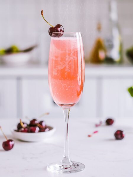 A Cherry Blossom Bellini served in a champagne flute with a soft pink hue, topped with bubbles and a fresh cherry garnish