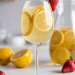 A glass of Limoncello Sangria with fresh strawberries and lemon slices, set against a bright, minimal white kitchen with a white countertop. A pitcher of sangria sits in the background, filled with the same golden sangria and floating citrus slices. The drink has a light, bubbly appearance, with condensation on the glass for a refreshing look.