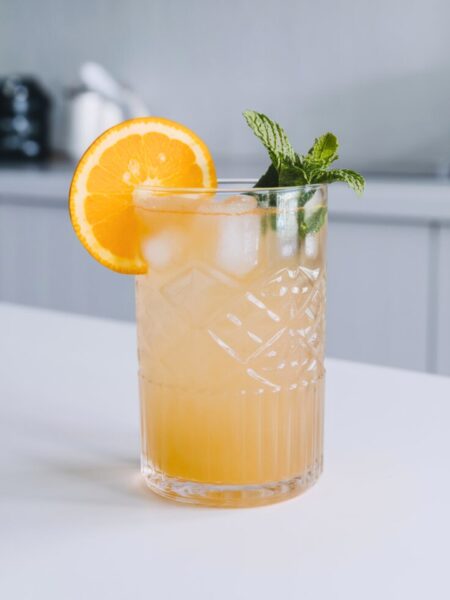 A refreshing glass of Coconut Citrus Refresher with ice, garnished with an orange slice and mint. The drink has a golden hue with a light, tropical glow.