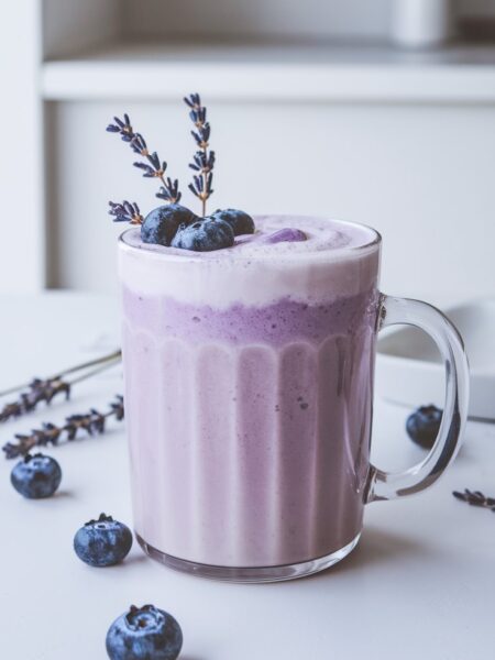 A warm, frothy Creamy Blueberry Lavender Latte served in a cozy mug with a soft purple hue. The drink is garnished with dried lavender and fresh blueberries, with steam rising for an inviting feel.