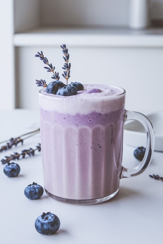 A warm, frothy Creamy Blueberry Lavender Latte served in a cozy mug with a soft purple hue. The drink is garnished with dried lavender and fresh blueberries, with steam rising for an inviting feel.