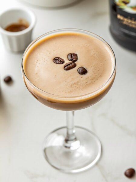 A Baileys Espresso Martini served in a sleek martini glass, garnished with three coffee beans. The drink has a rich, creamy color with a frothy top, set against a bright, minimal background with subtle coffee elements.