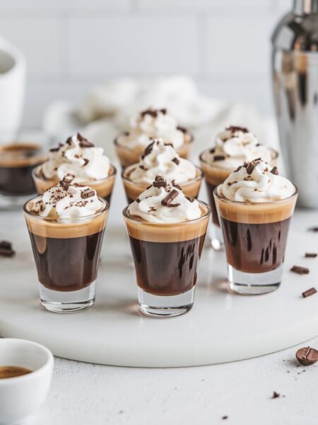 Espresso Martini Shots served in sleek shot glasses, topped with whipped cream and garnished with chocolate shavings. The drinks have a rich, dark base with a creamy, frothy topping, set against a bright, minimal background.