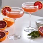 Two Blood Orange Lemon Drop Martinis served in elegant coupe glasses with sugar-rimmed edges, garnished with vibrant blood orange slices. The drinks have a rich reddish-orange hue, placed on a bright white countertop. Freshly sliced blood oranges, a cocktail shaker, and a small dish of sugar are arranged in the background, adding a stylish touch to the cocktail preparation scene.