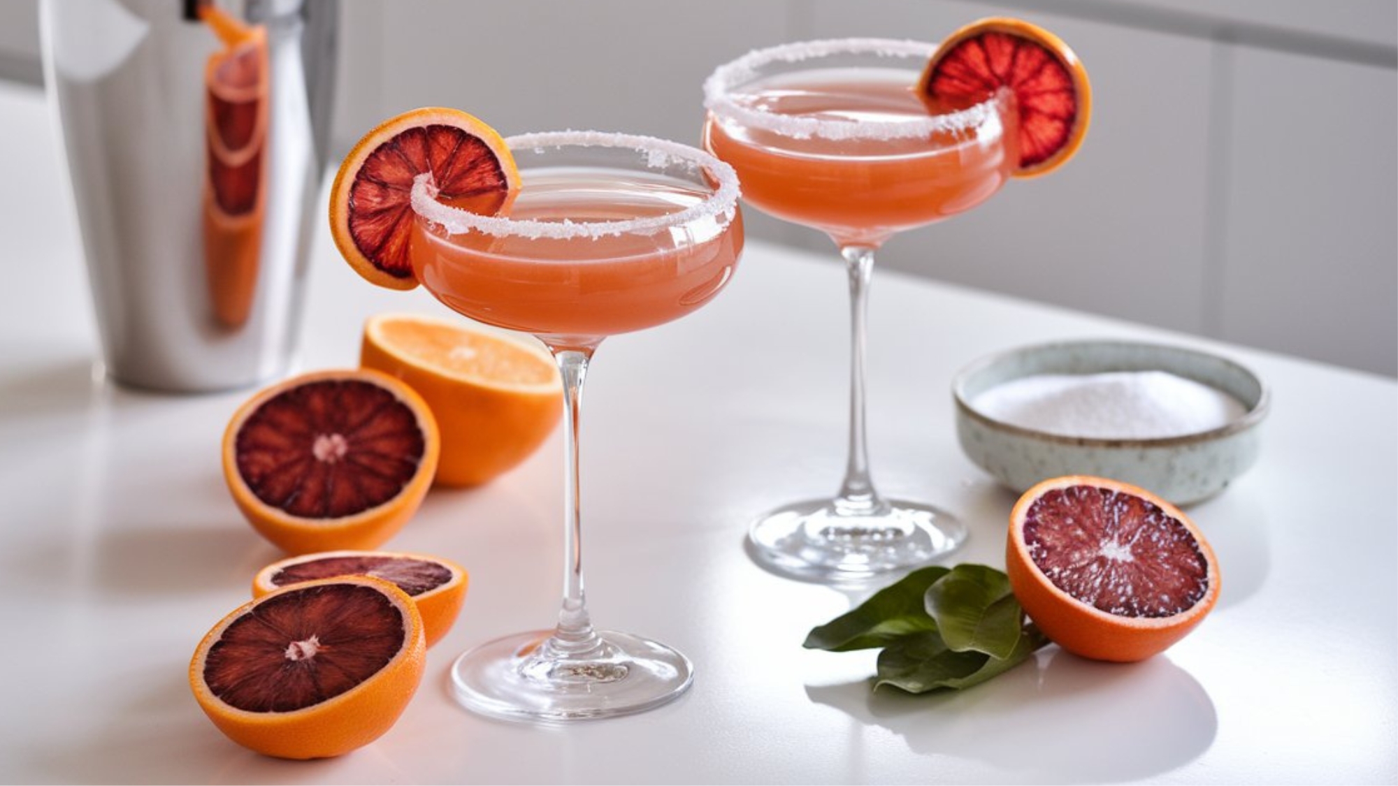 Two Blood Orange Lemon Drop Martinis served in elegant coupe glasses with sugar-rimmed edges, garnished with vibrant blood orange slices. The drinks have a rich reddish-orange hue, placed on a bright white countertop. Freshly sliced blood oranges, a cocktail shaker, and a small dish of sugar are arranged in the background, adding a stylish touch to the cocktail preparation scene.