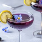 A vibrant Blueberry Limoncello Martini served in an elegant coupe glass, featuring a deep purple hue. The cocktail is garnished with a thin lemon slice on the rim, fresh blueberries, and a delicate edible blue flower floating on top. The background is a soft, neutral gray with scattered blueberries and lemon slices, creating a bright and refreshing spring-inspired setting.