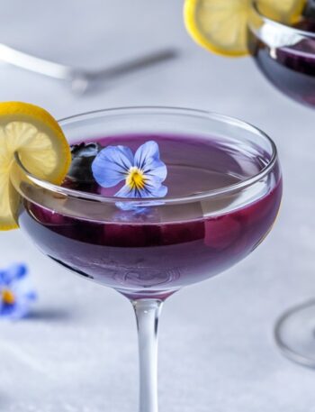 A vibrant Blueberry Limoncello Martini served in an elegant coupe glass, featuring a deep purple hue. The cocktail is garnished with a thin lemon slice on the rim, fresh blueberries, and a delicate edible blue flower floating on top. The background is a soft, neutral gray with scattered blueberries and lemon slices, creating a bright and refreshing spring-inspired setting.