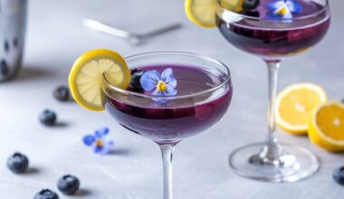 A vibrant Blueberry Limoncello Martini served in an elegant coupe glass, featuring a deep purple hue. The cocktail is garnished with a thin lemon slice on the rim, fresh blueberries, and a delicate edible blue flower floating on top. The background is a soft, neutral gray with scattered blueberries and lemon slices, creating a bright and refreshing spring-inspired setting.