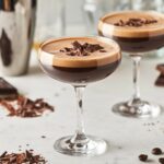 Two Chocolate Espresso Martinis served in coupe glasses on a white marble countertop. Each martini has a rich, dark coffee hue with a thick, velvety crema on top and is garnished with chocolate shavings. In the softly blurred background, a stainless steel cocktail shaker, dark chocolate bars, cocoa powder, and coffee beans enhance the indulgent cocktail aesthetic.