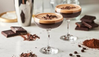 Two Chocolate Espresso Martinis served in coupe glasses on a white marble countertop. Each martini has a rich, dark coffee hue with a thick, velvety crema on top and is garnished with chocolate shavings. In the softly blurred background, a stainless steel cocktail shaker, dark chocolate bars, cocoa powder, and coffee beans enhance the indulgent cocktail aesthetic.