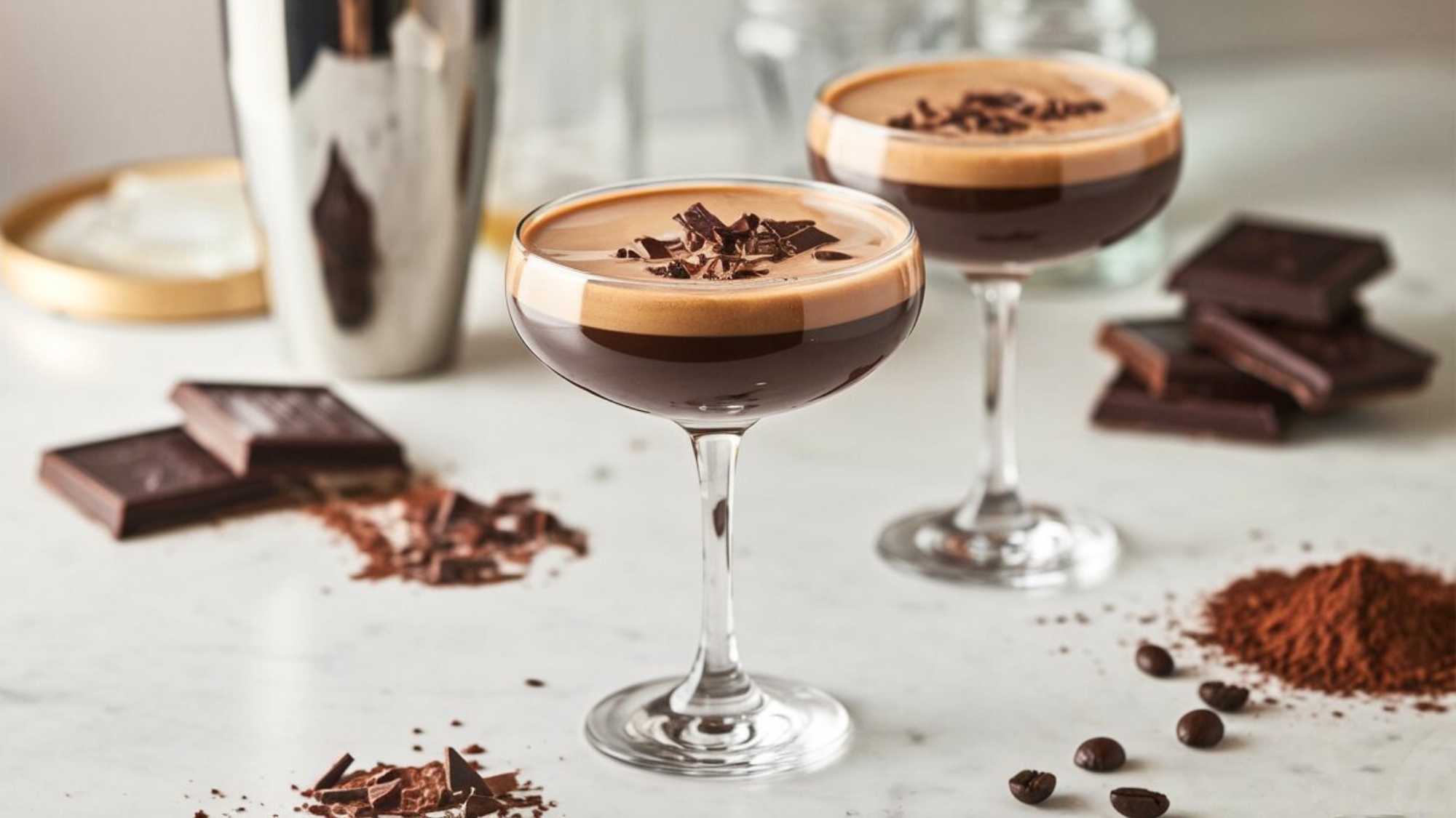 Two Chocolate Espresso Martinis served in coupe glasses on a white marble countertop. Each martini has a rich, dark coffee hue with a thick, velvety crema on top and is garnished with chocolate shavings. In the softly blurred background, a stainless steel cocktail shaker, dark chocolate bars, cocoa powder, and coffee beans enhance the indulgent cocktail aesthetic.