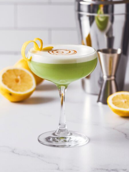 A vibrant green Leprechaun Sour cocktail in a coupe glass with a frothy egg white top, garnished with a lemon twist and a few drops of Angostura bitters for decoration.