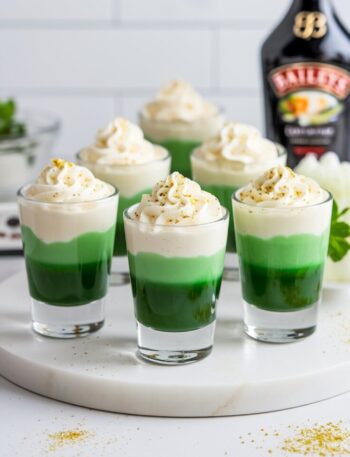 A set of vibrant Lucky Leprechaun Shots served in clear shot glasses, featuring a clean green and cream layered effect, topped with whipped cream and gold sprinkles.